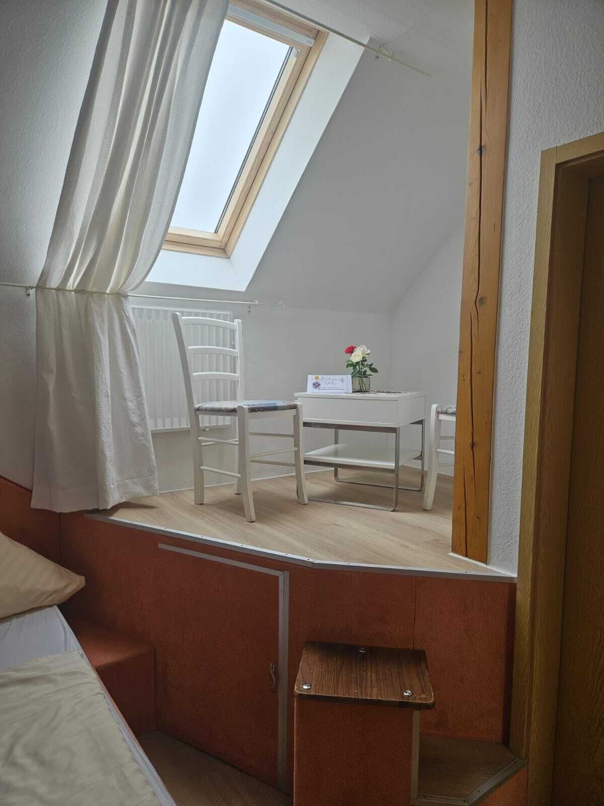 Een kleine, schuine kamer met een dakraam, een witte stoel, een tafeltje met een vaas en een bed.