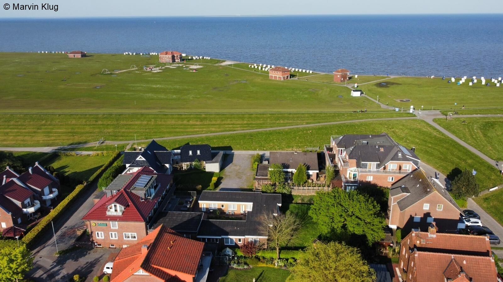 An aerial view of a residential area with houses, a grassy field, and a body of water in the background.