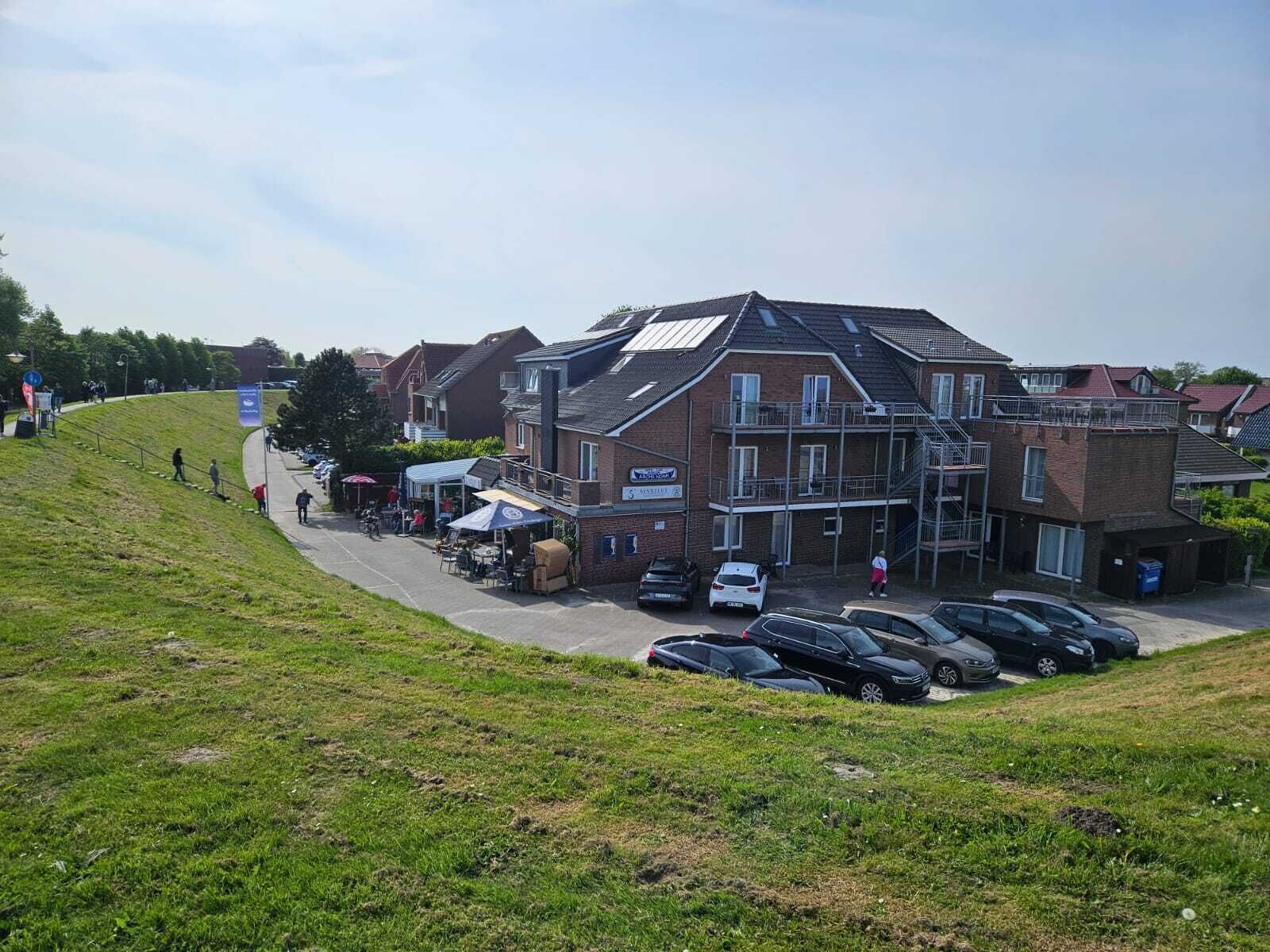 Een meerlaags gebouw met een parkeerplaats ervoor, een grasheuvel en enkele mensen die lopen.