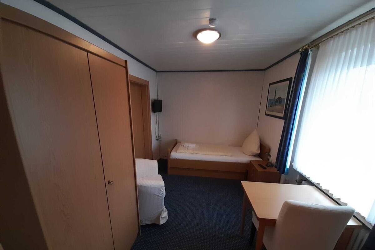 A compact room with a bed, a desk, a chair, a wardrobe, and a window with curtains.