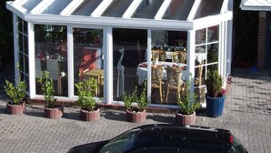 A conservatory with a glass roof, surrounded by potted plants, and a car parked in front. - Hotel Arche Noah (Wangerland)