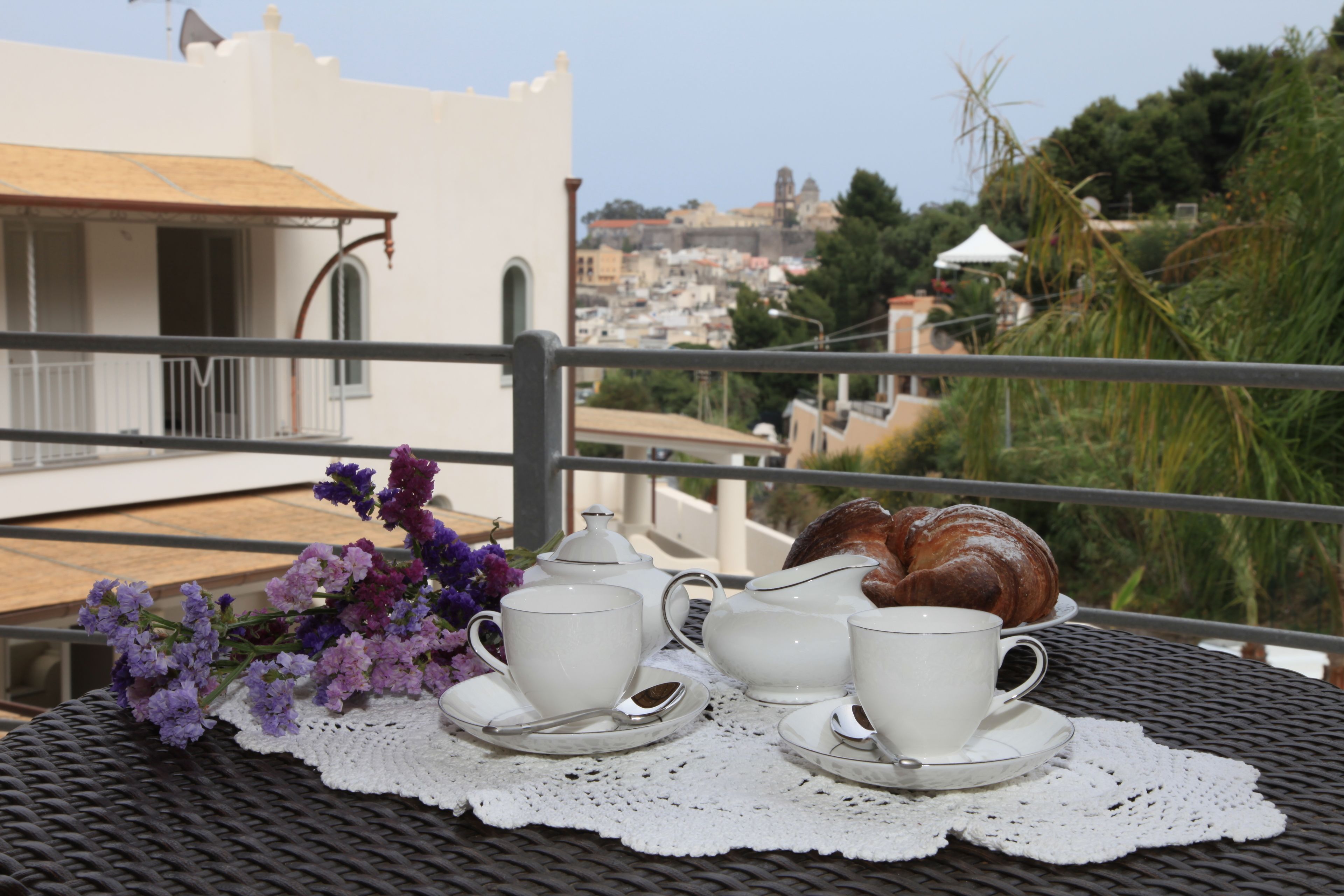 Una mesa con tazas blancas, platillos y una tetera sobre un encaje, junto con un croissant y flores moradas, en un balcón con vistas a un paisaje urbano.