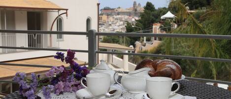 Una mesa con tazas blancas, platillos y una tetera sobre un encaje, junto con un croissant y flores moradas, en un balcón con vistas a un paisaje urbano.