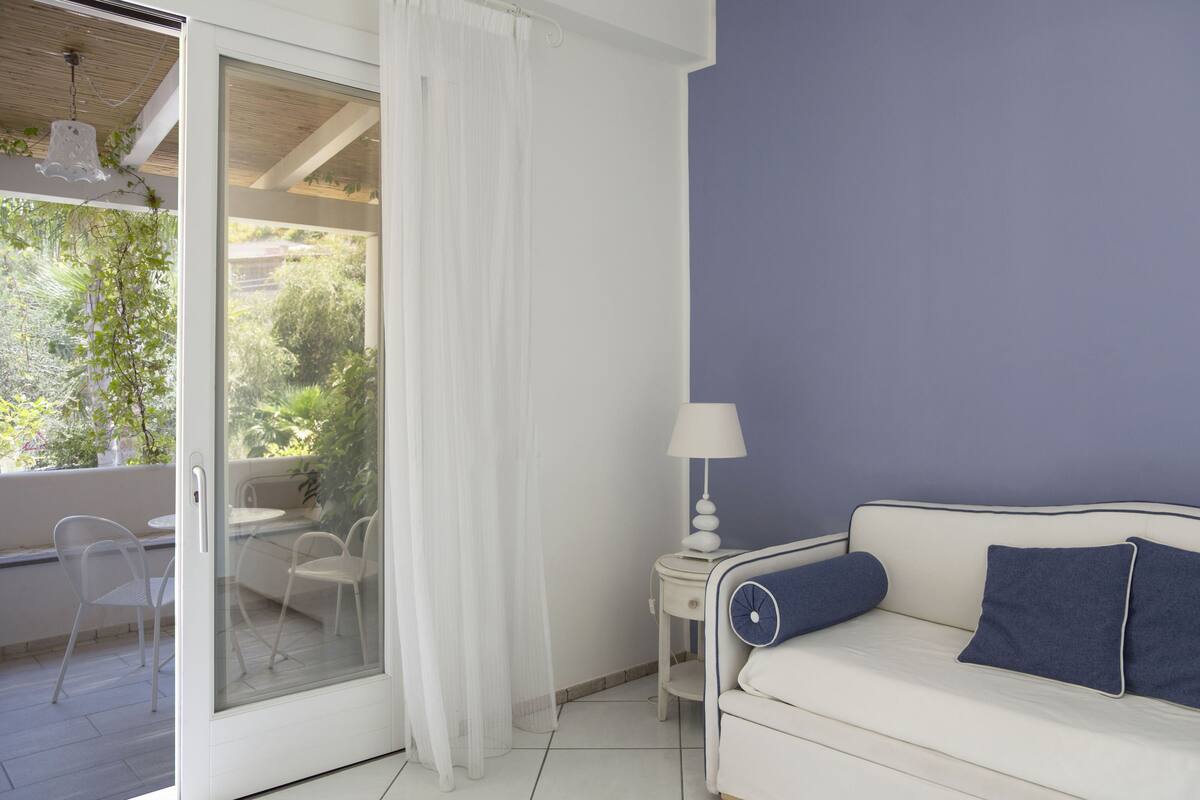 Ein Zimmer mit einem weißen Sofa, einem kleinen Beistelltisch mit einer Lampe und einer Glastür, die zu einem Außenbereich mit Sitzgelegenheiten führt.