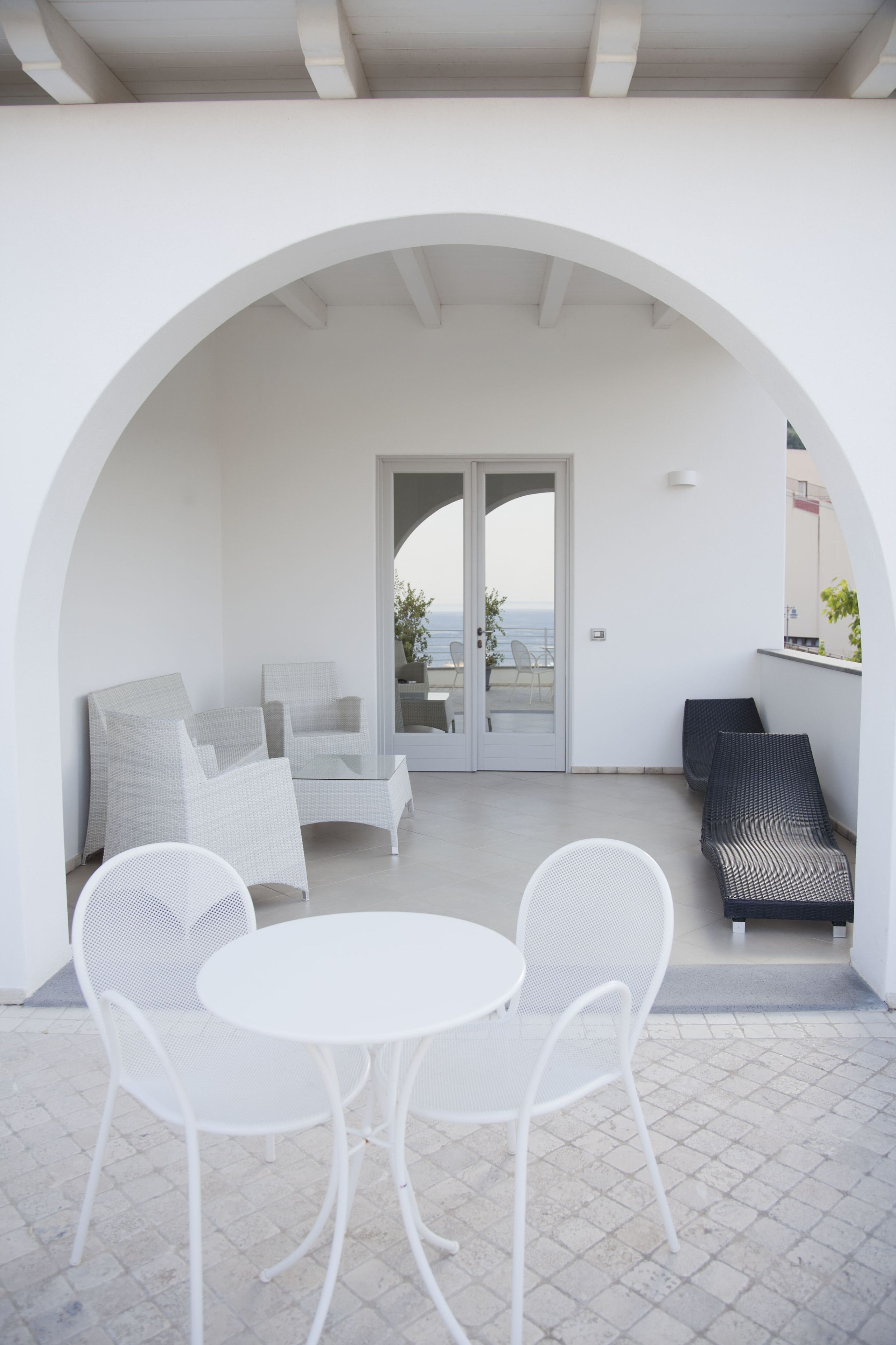 A modern outdoor seating area with white chairs and a table, a glass door leading to a balcony, and a view of the sea.