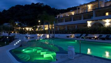 A swimming pool with a green-lit jacuzzi area, lounge chairs, and a building with multiple balconies in the background.