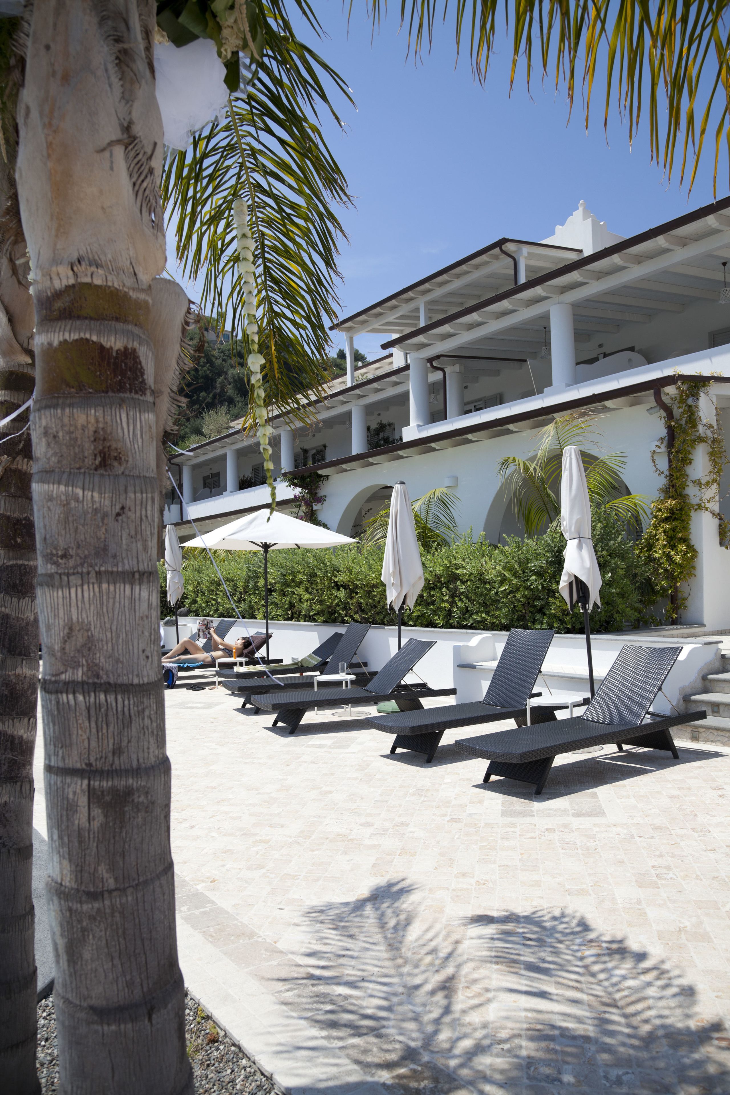 Un área de piscina con sillones de descanso y sombrillas, palmeras y un edificio de fachada blanca y arcos.