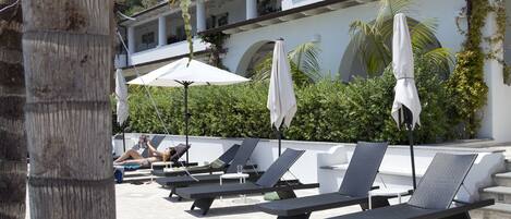 Un área de piscina con sillones de descanso y sombrillas, palmeras y un edificio de fachada blanca y arcos.