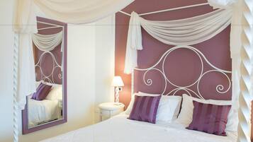 Un letto ben rifatto con lenzuola bianche e cuscini viola, un comodino con una lampada e uno specchio con cornice decorativa.