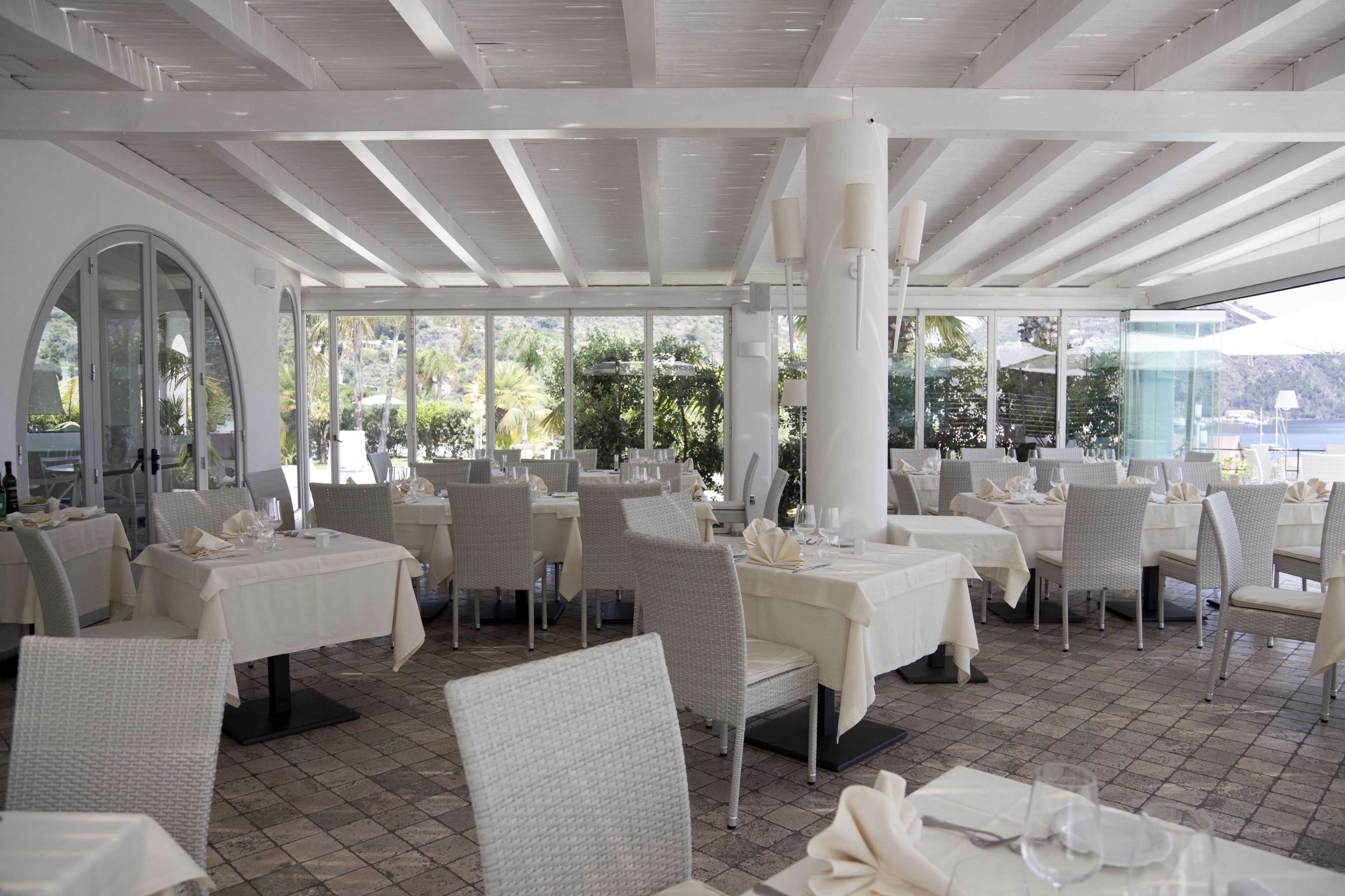 Un ristorante con tavoli e sedie bianche, ampie finestre e vista su spazi verdi e sull'acqua.