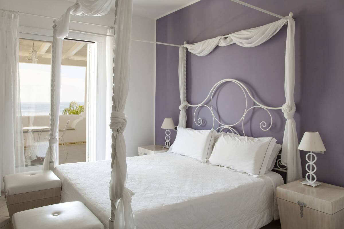 Ein Bett mit Himmelbett, ausgestattet mit weißer Bettwäsche, ein Nachttisch mit Lampe und Blick ins Freie durch eine Glastür.