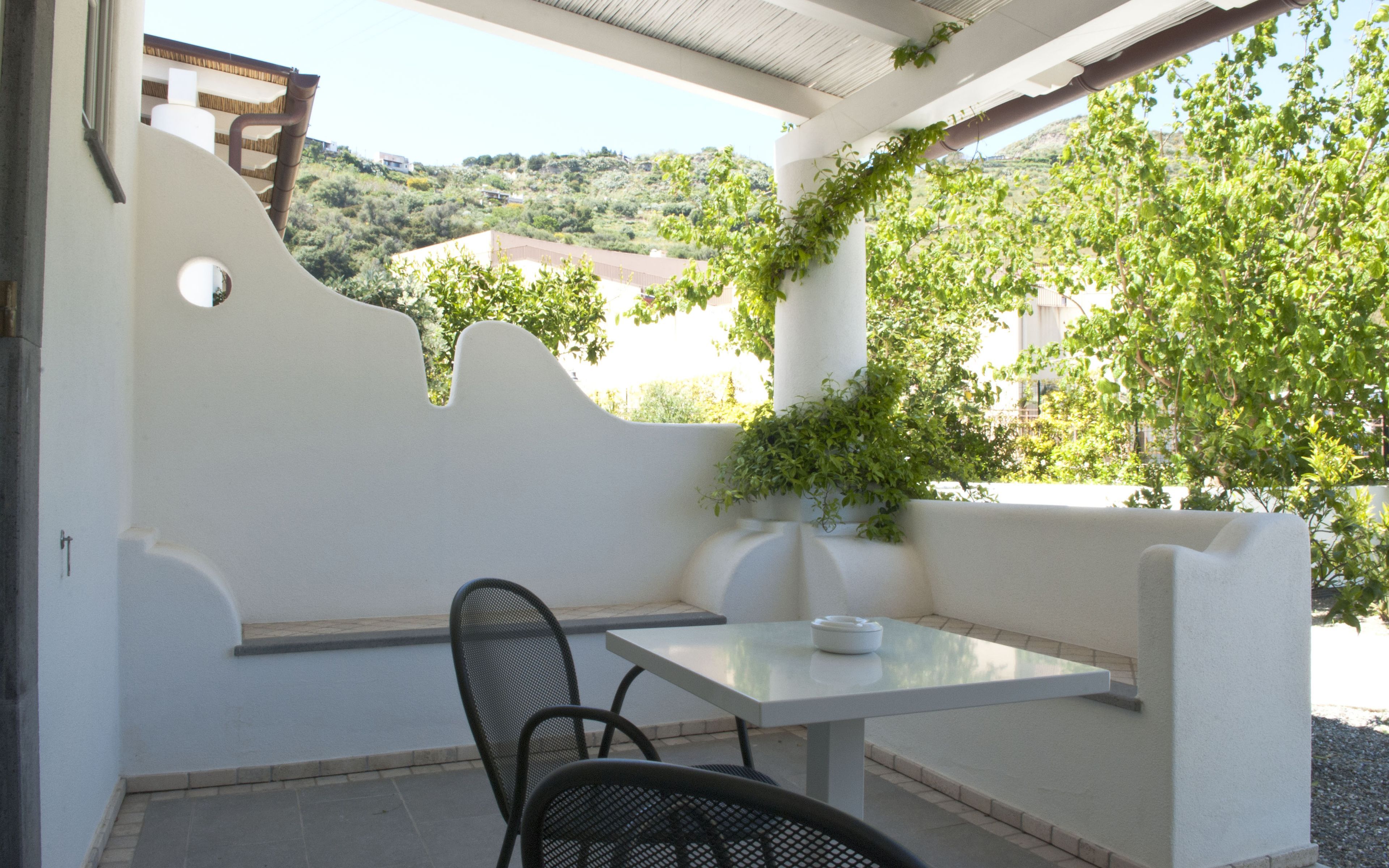 An outdoor patio with a white table and black metal chairs, surrounded by white walls and a view of a hillside with buildings.