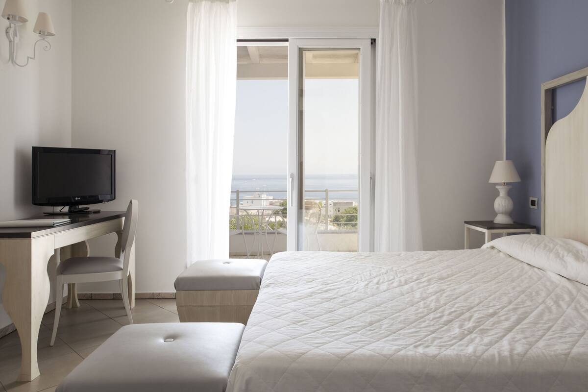 Ein Hotelzimmer mit Bett, Schreibtisch mit Fernseher, Stuhl, Nachttisch mit Lampe und einem Balkon mit Aussicht.