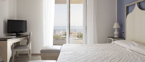A hotel room with a bed, a desk with a TV, a chair, a nightstand with a lamp, and a balcony with a view.