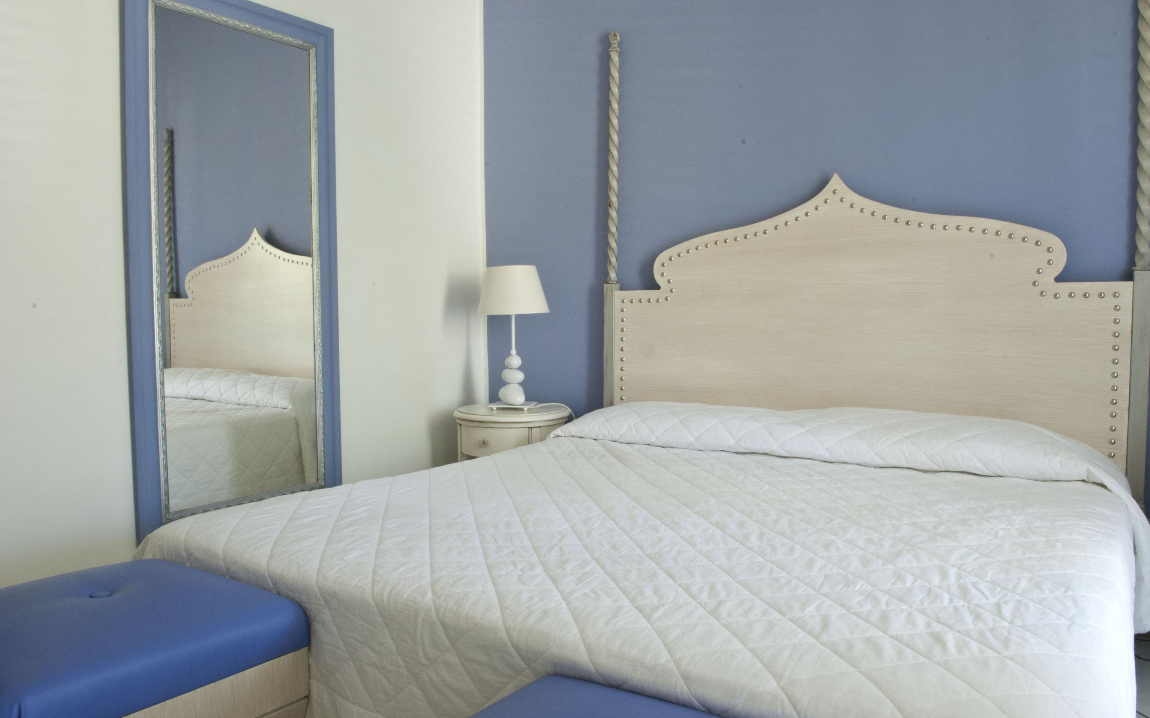 Una cama bien hecha con un edredón blanco acolchado, una mesita de noche con una lámpara y un espejo en la pared.