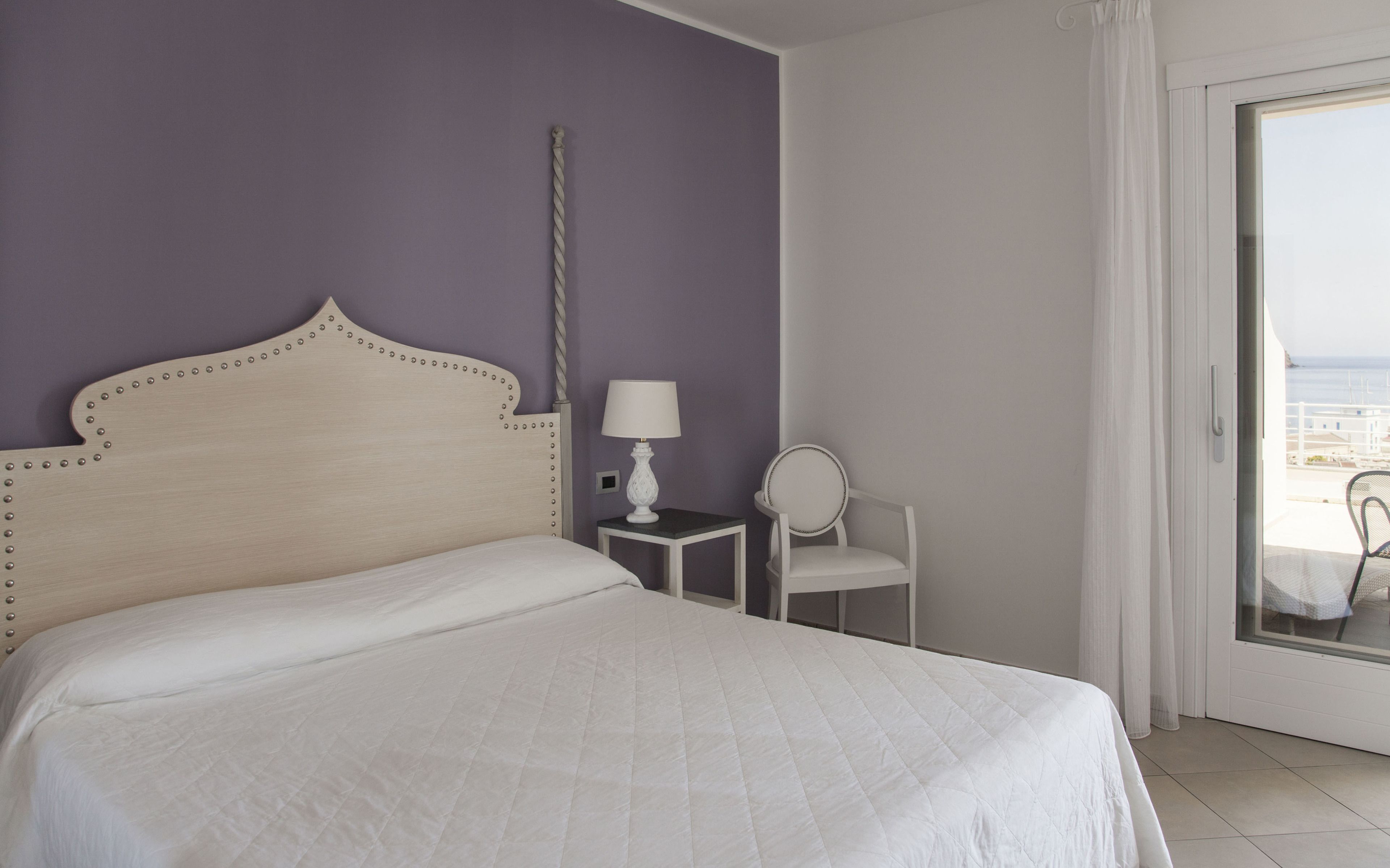 A bedroom with a bed, a bedside table with a lamp, a chair, and a view of the sea through a glass door.