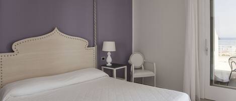 A bedroom with a bed, a bedside table with a lamp, a chair, and a view of the sea through a glass door.