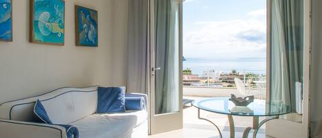 Quarto com sofá branco, mesa de vidro e vista para o mar.