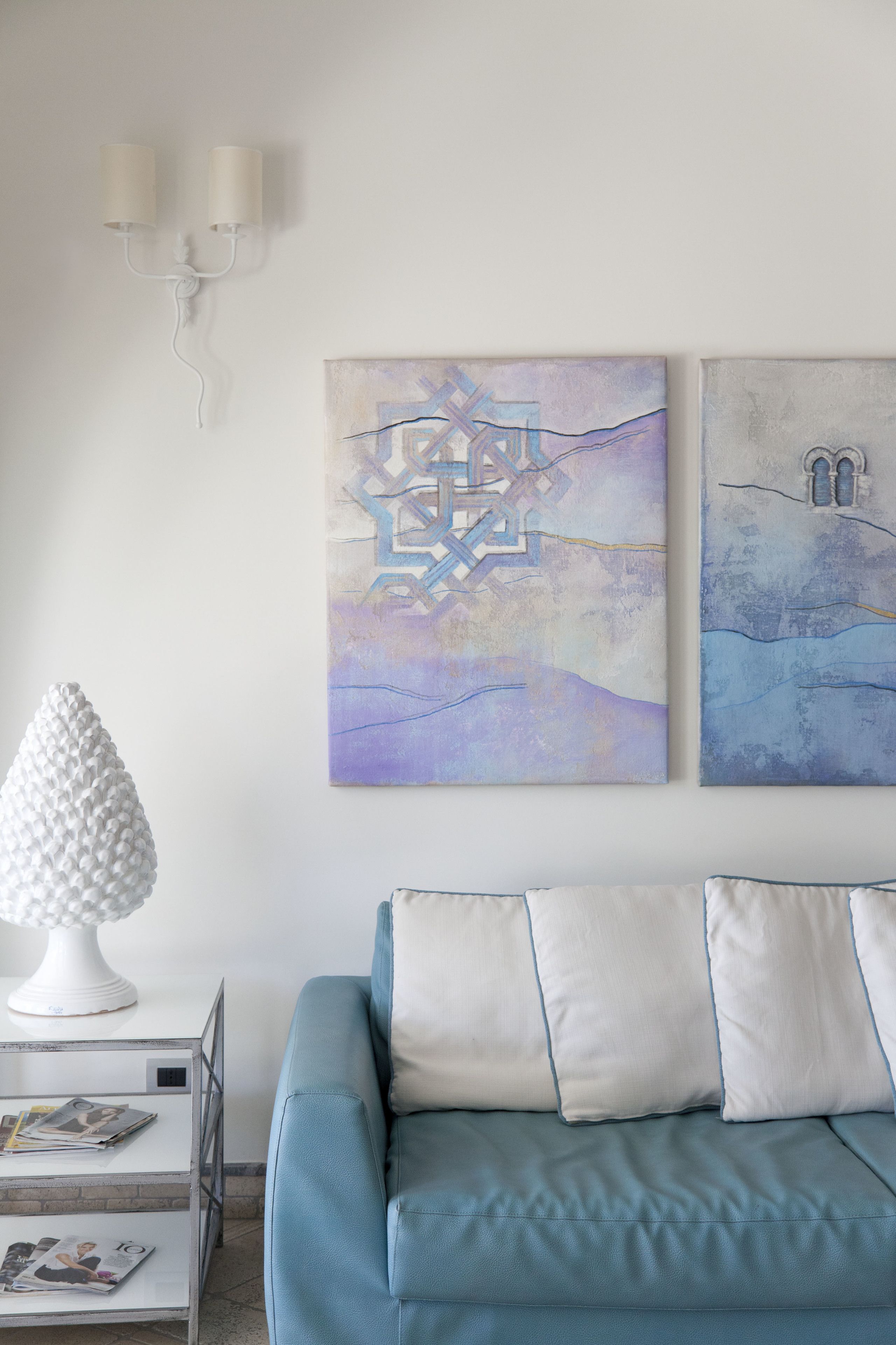 A modern living room with a blue sofa, white cushions, a white lamp, and two abstract paintings on the wall.