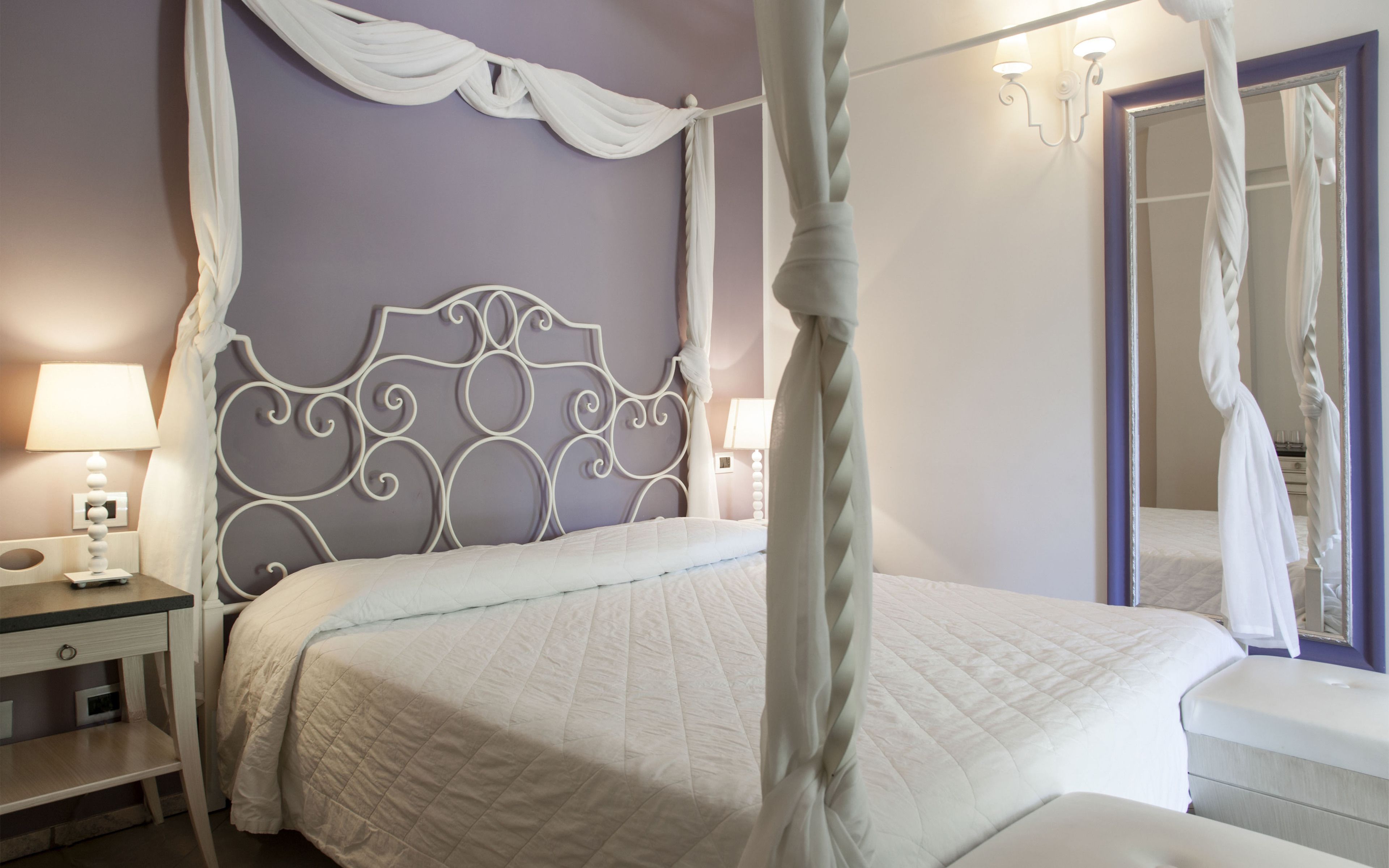 A four-poster canopy bed with a white headboard, bedside tables with lamps, and a mirror on the wall.