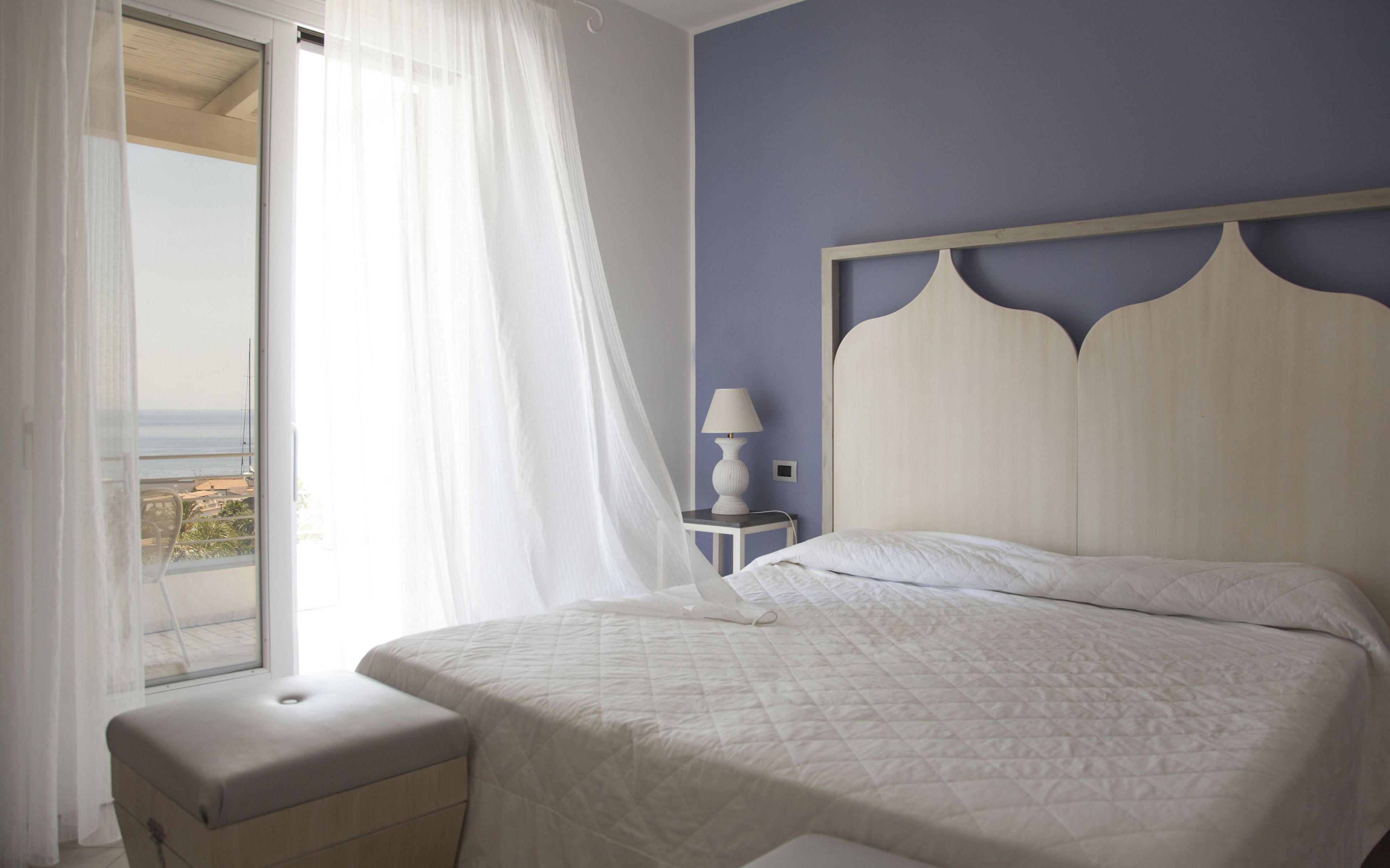 Un dormitorio con cama, mesita de noche con lámpara, ventana con cortinas translúcidas y vista al mar.