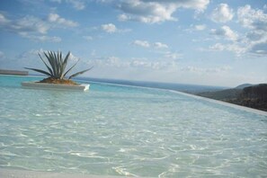 Vista despejada de una piscina con una isla de plantas en el centro, rodeada por un cielo azul brillante y colinas lejanas.