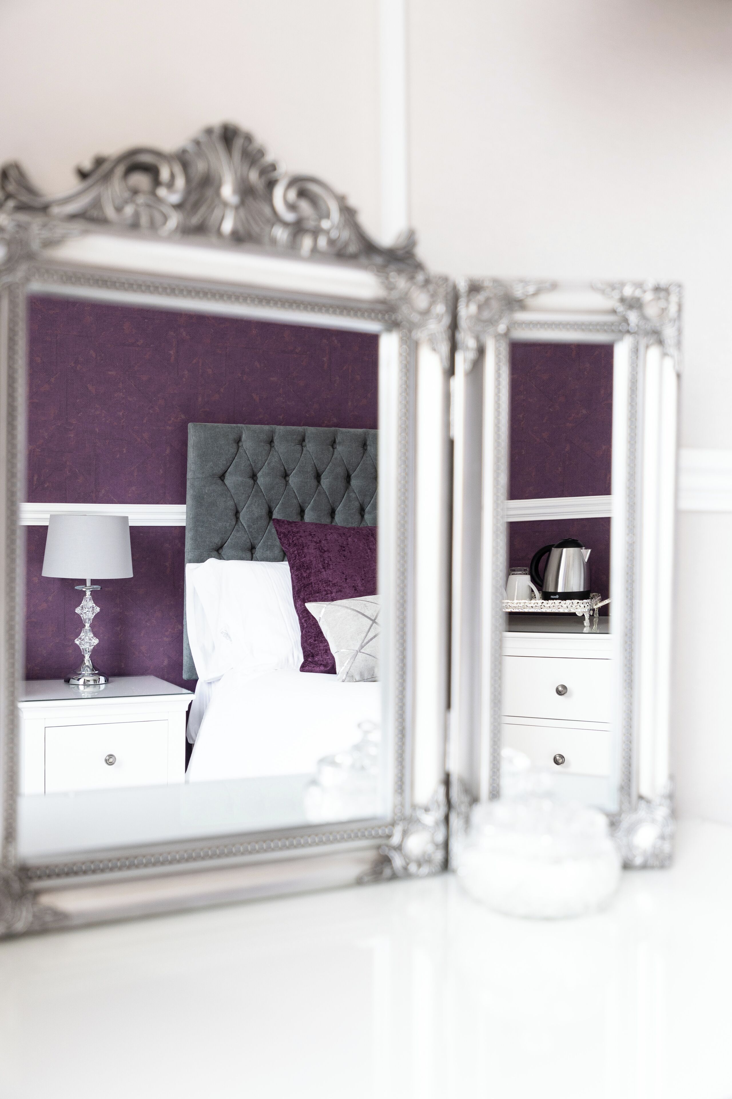 A bedroom with a tufted headboard, a white nightstand, a lamp, and a bedside table with a decorative item.