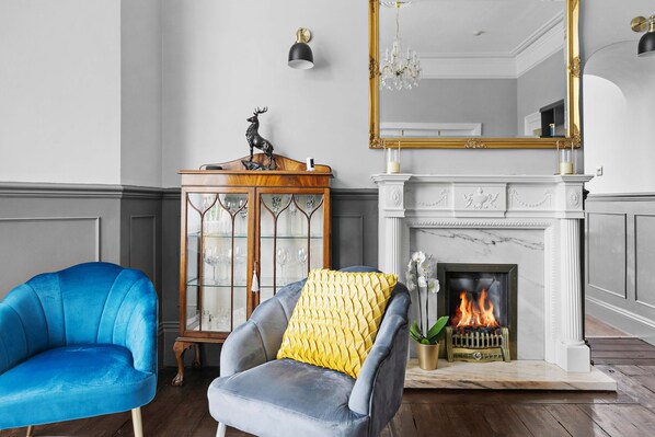 A room with a blue armchair, a gray armchair with a yellow pillow, a wooden side table with a deer statue, a glass cabinet, a fireplace, and a large mirror with a gold frame. - The Berburry (Torquay)