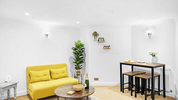 A living room with a yellow sofa, a round coffee table, and a small dining table with chairs. There is a potted plant and a vase with flowers on the dining table.