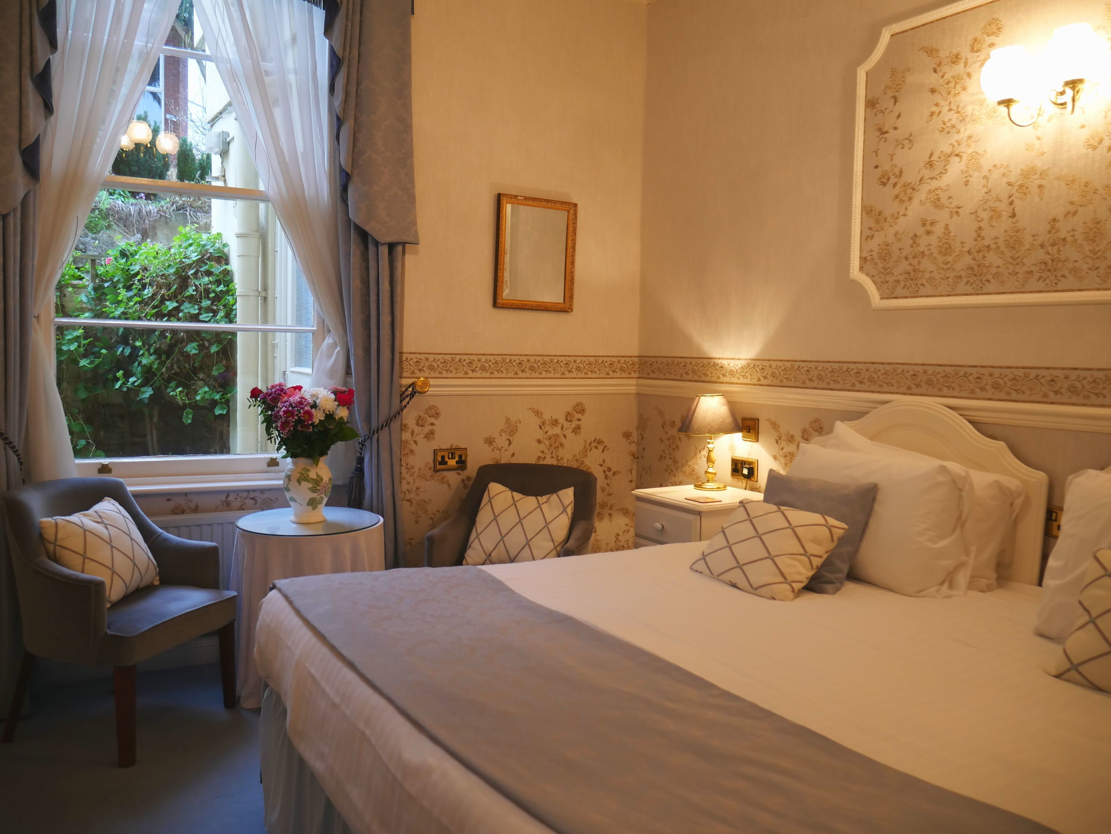 Una habitación de hotel con una cama, una silla, una mesita con un jarrón de flores, una mesita de noche con una lámpara y una ventana con cortinas.