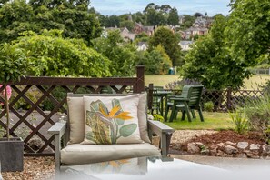 A garden seating area with a cushioned chair and a table, surrounded by greenery and a view of houses in the distance. - The Berburry (Torquay)