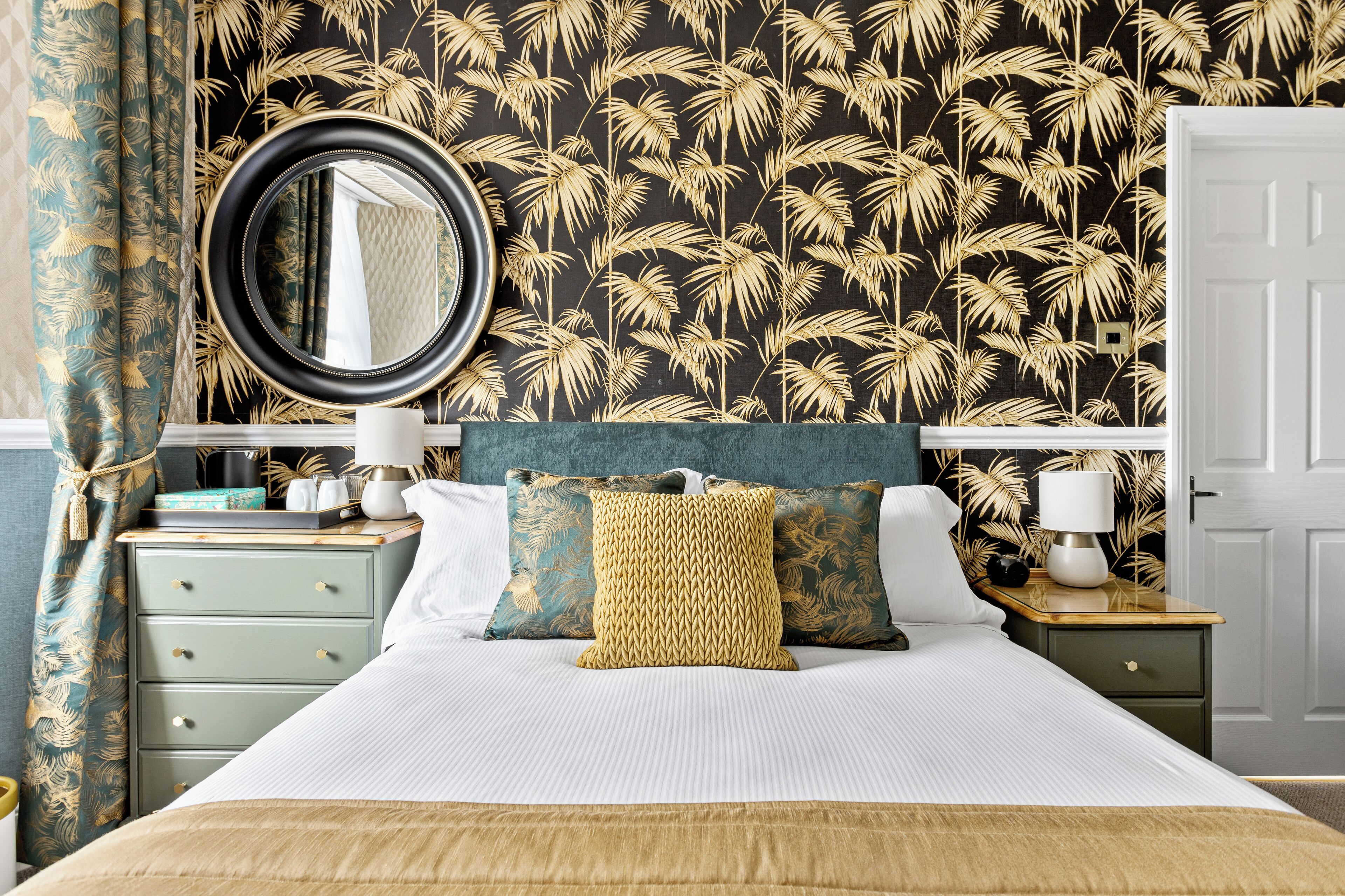 A neatly made bed with a white comforter and two pillows, a bedside table with a lamp, and a wall with a palm leaf pattern.