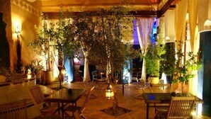 Un patio interior con mesas y sillas, rodeado de plantas en macetas e iluminación decorativa.