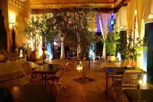 Un patio interior con mesas y sillas, rodeado de plantas en macetas e iluminación decorativa.