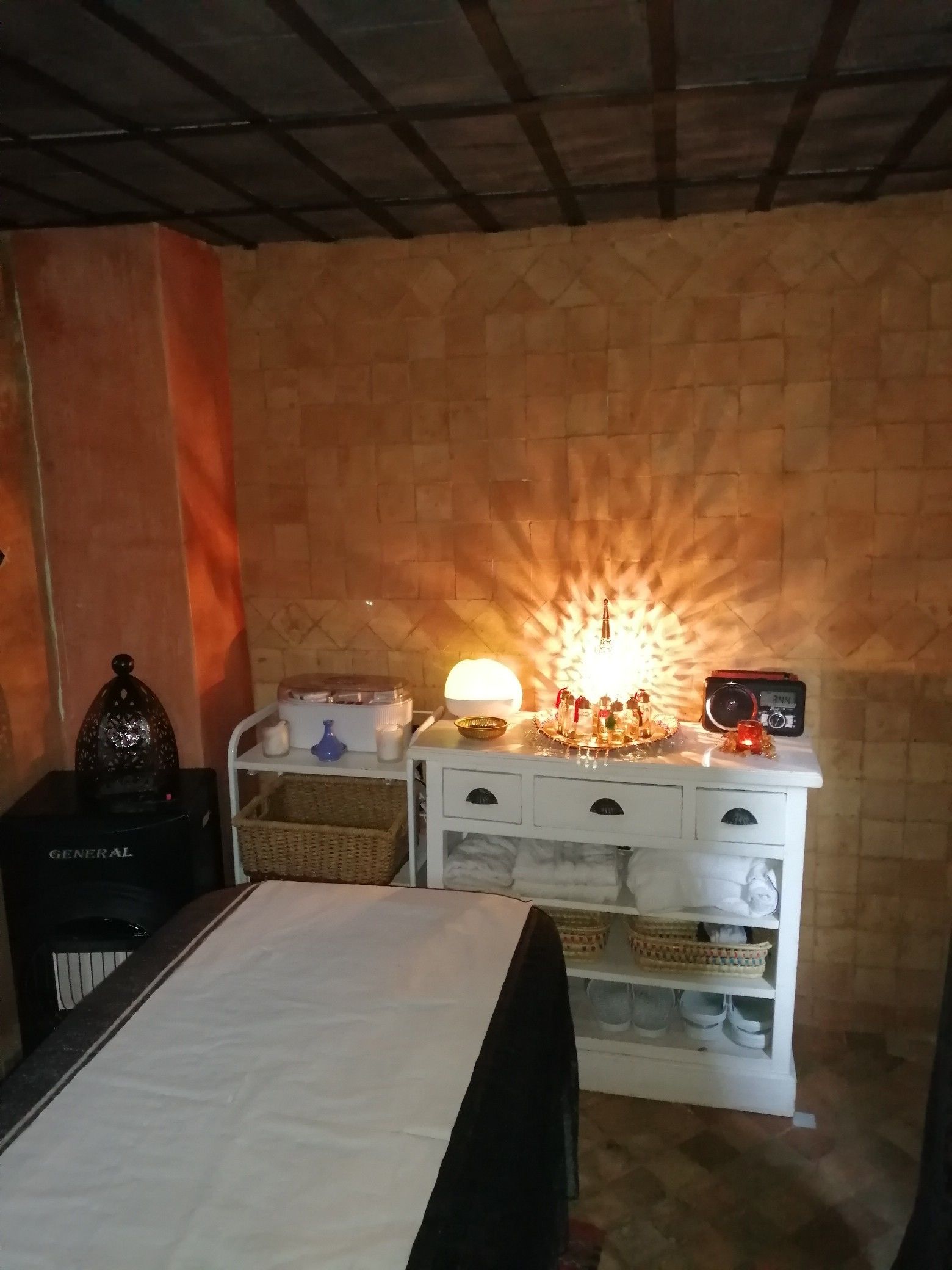 Una habitación con una cama, un aparador blanco con cajones, una radio, una lámpara y un objeto decorativo en el aparador.