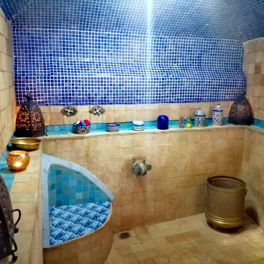 A bathroom with a blue-tiled shower area, a sink with a blue patterned basin, and a shelf with various toiletries.