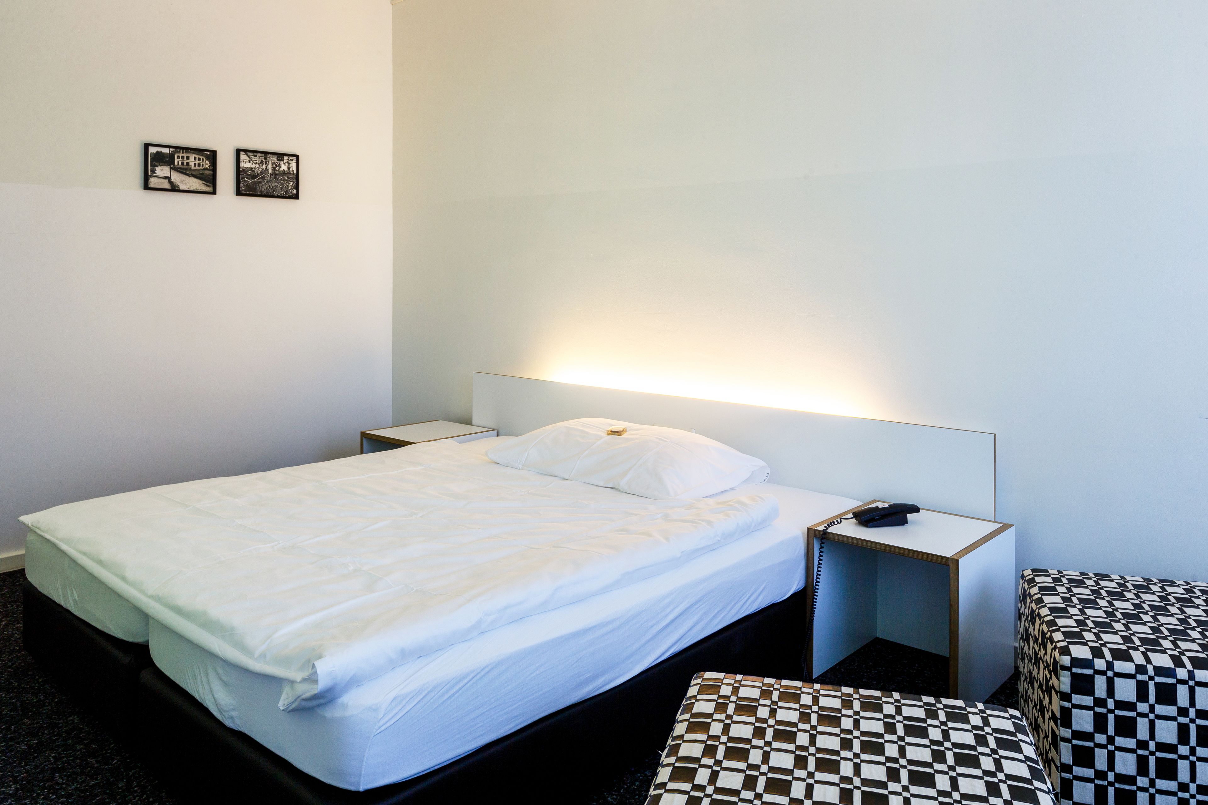 A hotel room with a single bed, a bedside table with a phone, a small bedside table with a lamp, a checkered bench, and two framed pictures on the wall.