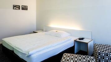 A hotel room with a single bed, a bedside table with a phone, a small bedside table with a lamp, a checkered bench, and two framed pictures on the wall.