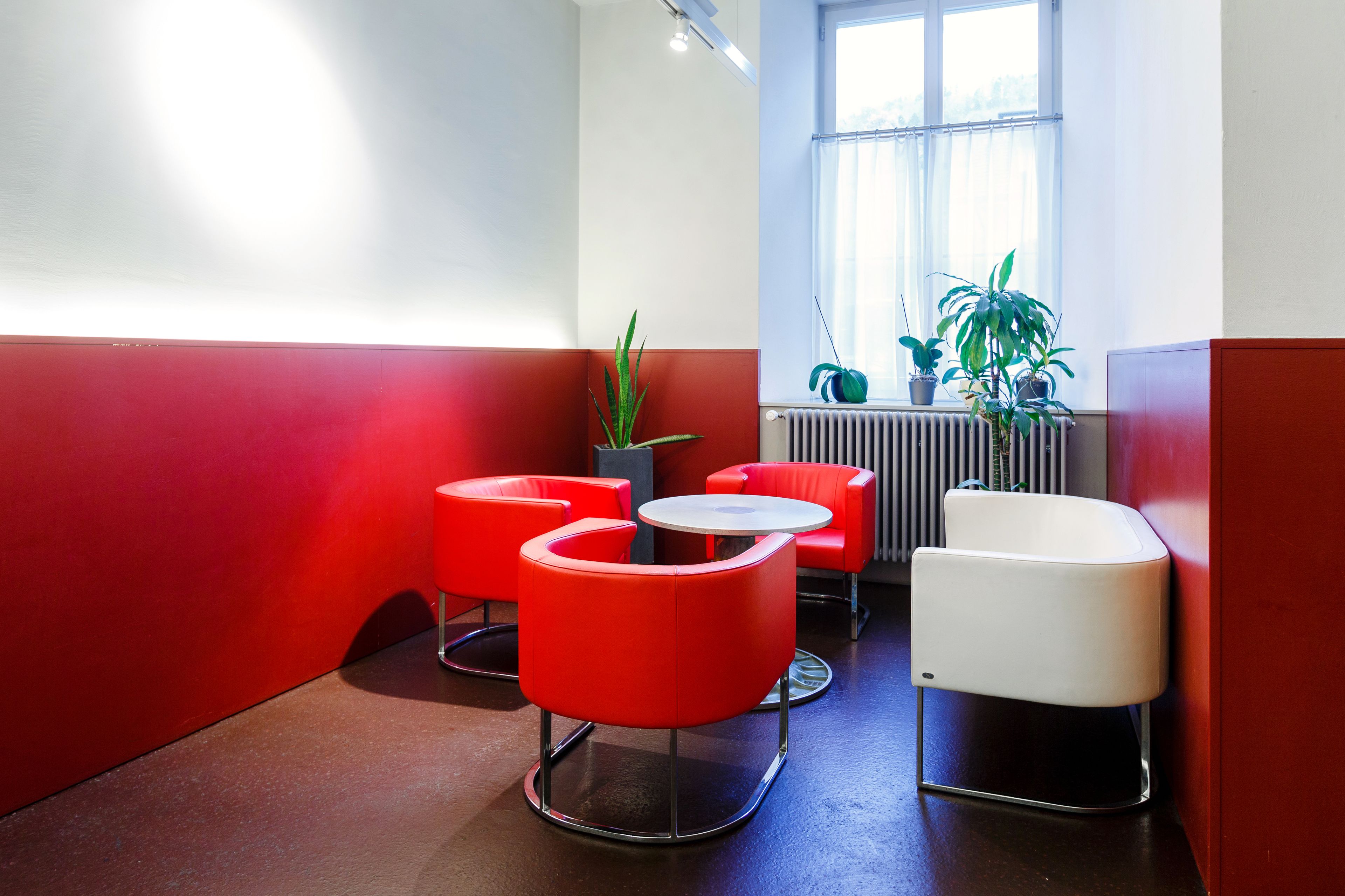 A room with red and white walls, a radiator, a window with plants, and red and white chairs.