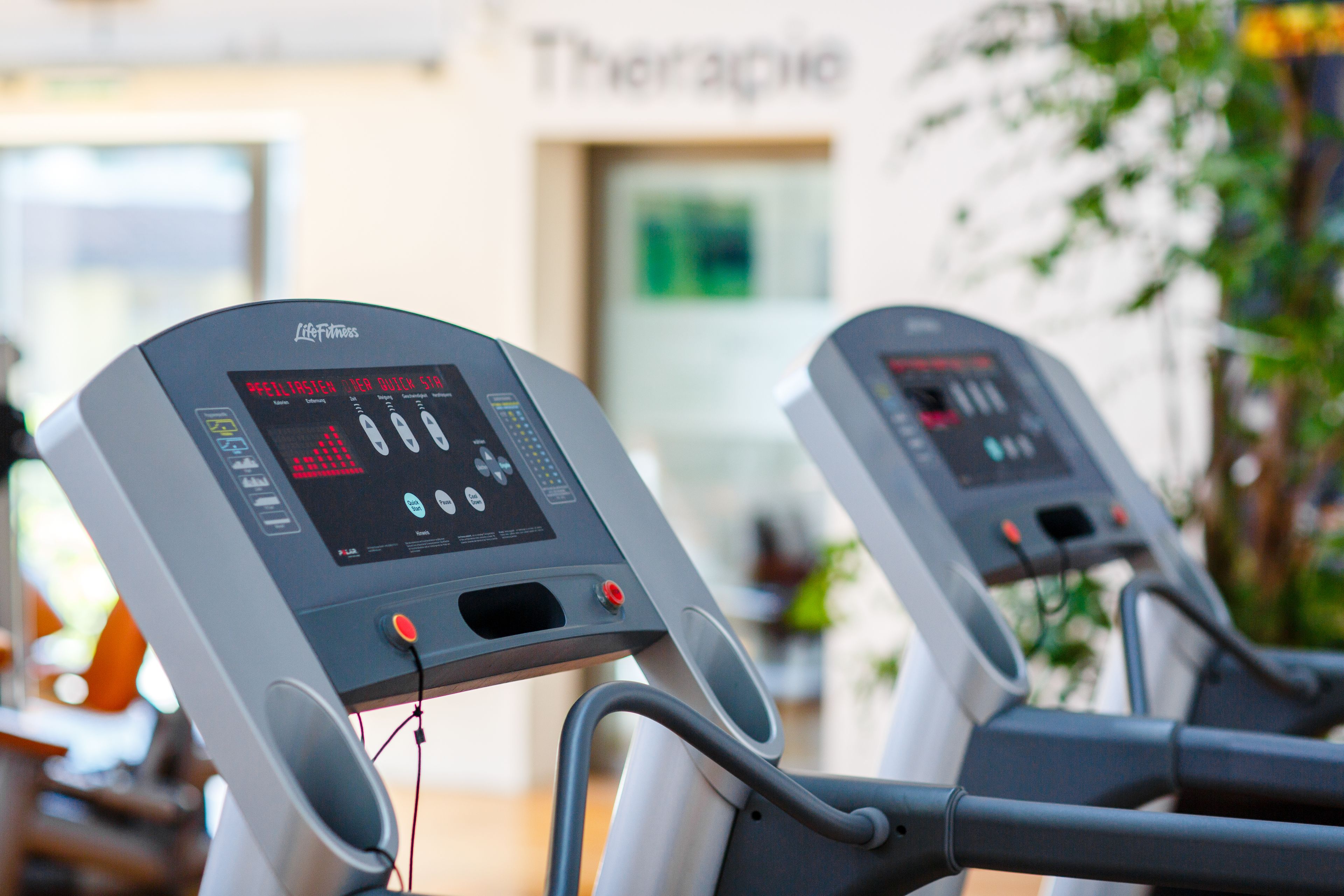 Zwei Trainingsgeräte mit digitalen Anzeigen in einem Raum, auf dessen Schild „Therapie“ steht.