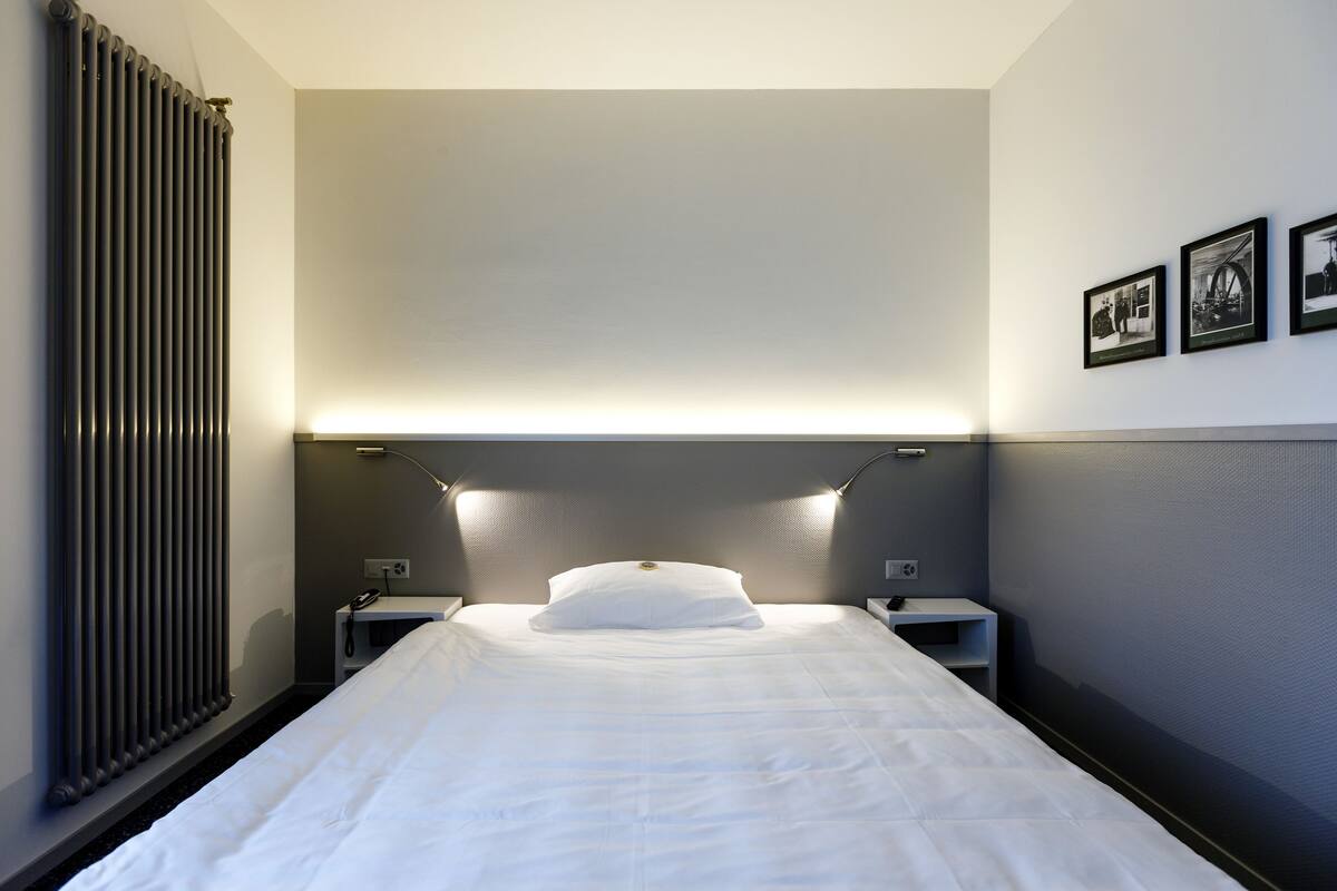A hotel room with a neatly made bed, a radiator, a nightstand with a lamp, and three framed pictures on the wall.