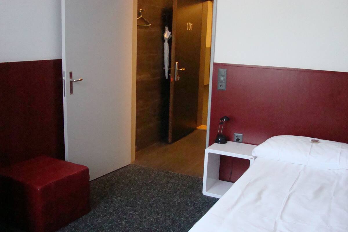 A hotel room with a bed, a small bedside table, a red chair, and a door leading to a bathroom.