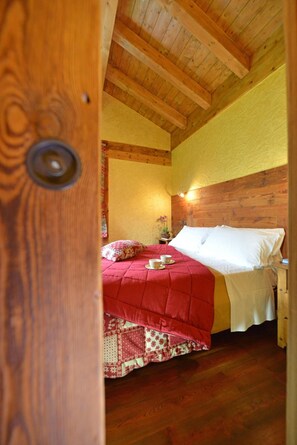 A cozy bedroom with a wooden ceiling, a red bedspread, and a nightstand with a lamp. - Le Petit Abri (Ayas)