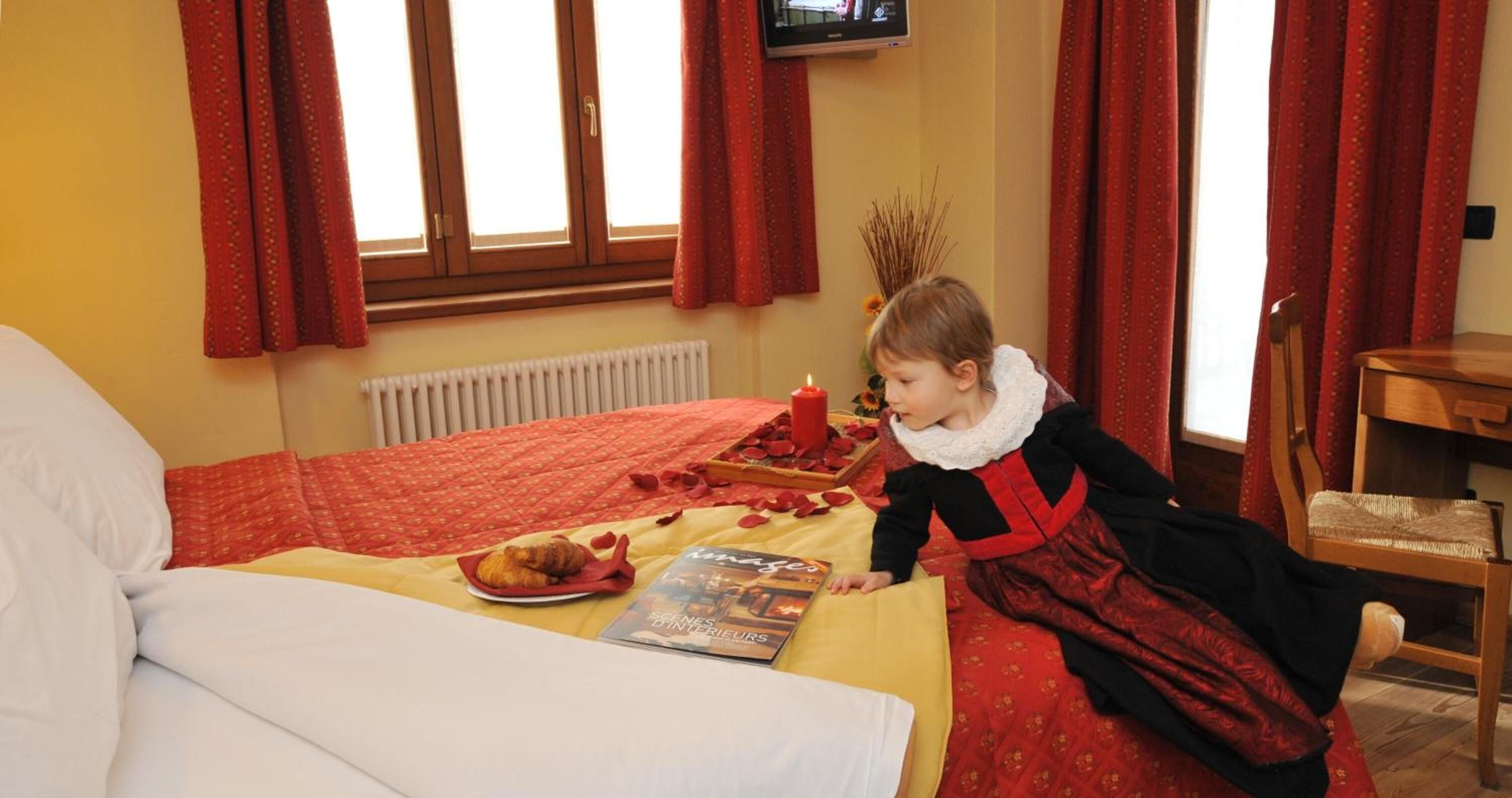A child in a red and black dress sitting on a bed with a red blanket, a book, and a plate with food.