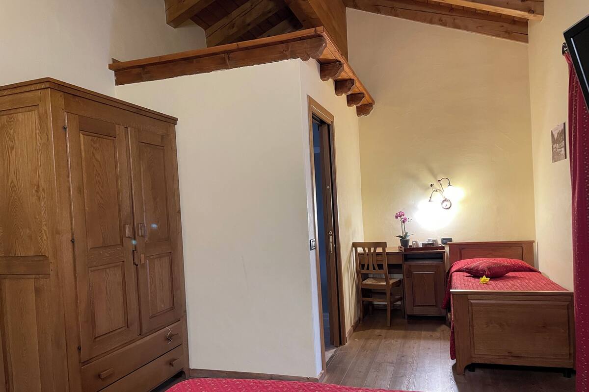 Un dormitorio con un armario de madera, una cama con colcha roja, un escritorio con silla y una mesita de noche con lámpara.
