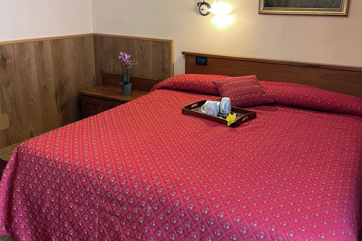 Una cama bien hecha con colcha roja, una mesita de noche con lámpara y un cuadro en la pared.