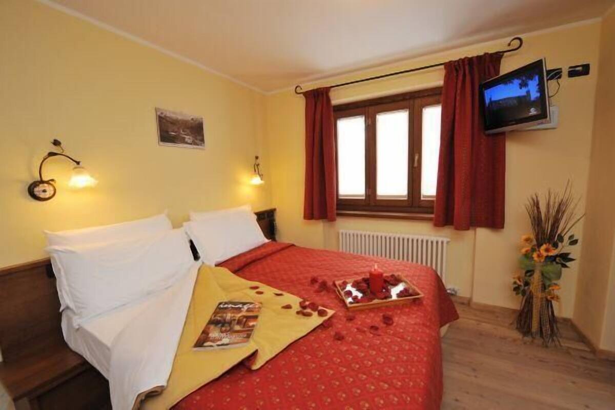 Una habitación de hotel con una cama, televisión, ventana con cortinas y una lámpara fijada a la pared.