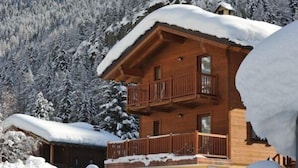 Uma cabana de madeira com varanda, rodeada por árvores e montanhas cobertas de neve.