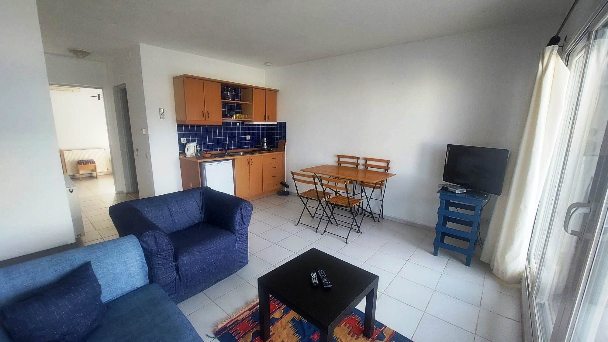 Un espacio habitable compacto con sofá, mesa de comedor, sillas, una pequeña cocina y televisor sobre un mueble.