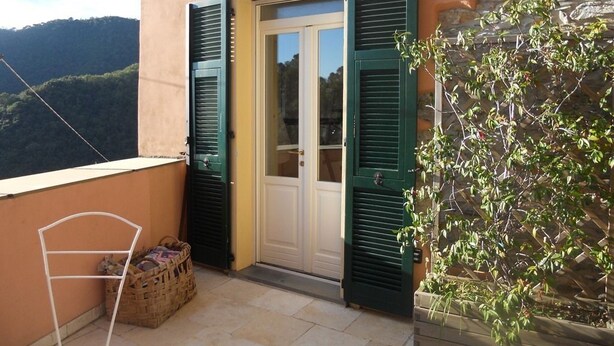 Un balcón con una cesta de mimbre, una puerta con contraventanas verdes y una pared de piedra con plantas trepadoras.